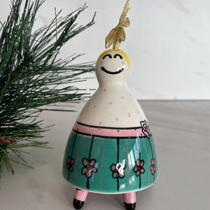 May include: A ceramic figurine with a smiling face, blonde hair, and a gold glitter accent. The figurine has a white body with pink polka dots, a pink band, and a teal skirt adorned with pink flowers. It stands on pink legs with black feet.