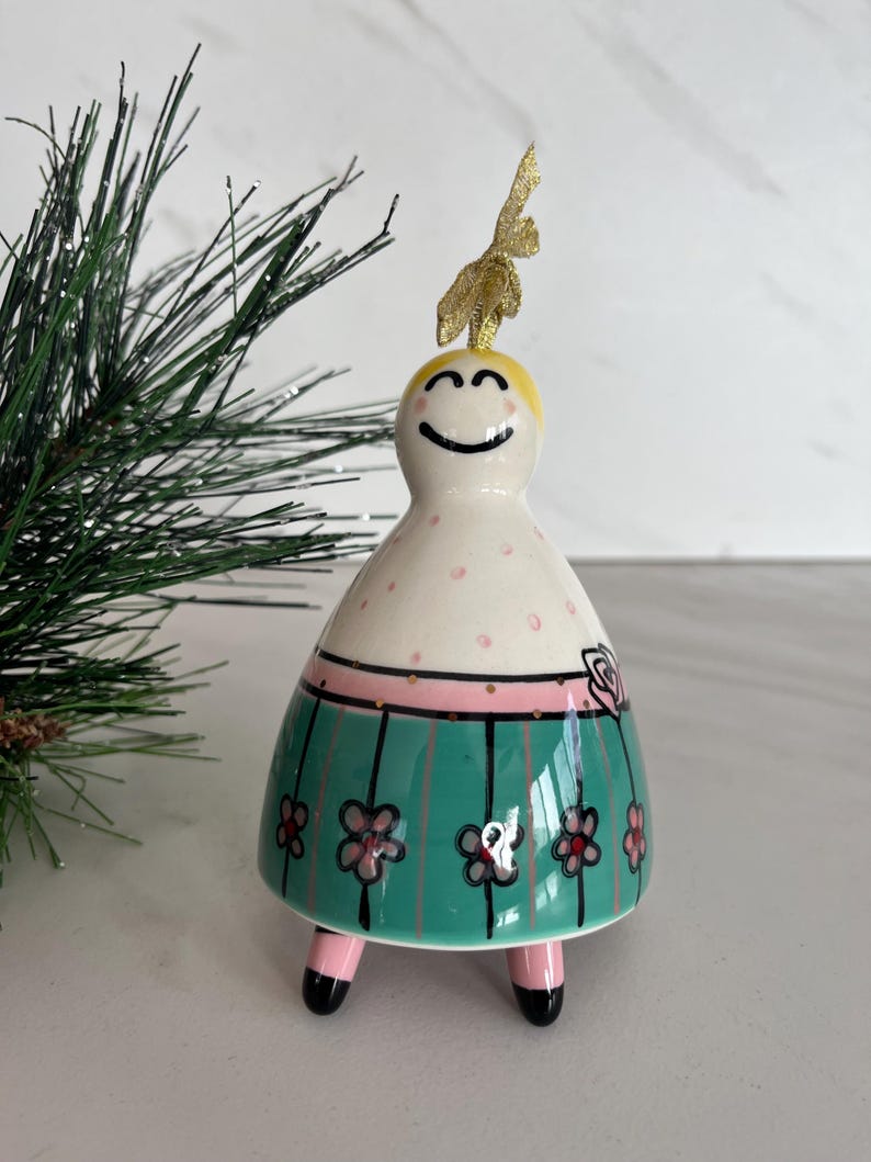 May include: A ceramic figurine with a smiling face, blonde hair, and a gold glitter accent. The figurine has a white body with pink polka dots, a pink band, and a teal skirt adorned with pink flowers. It stands on pink legs with black feet.