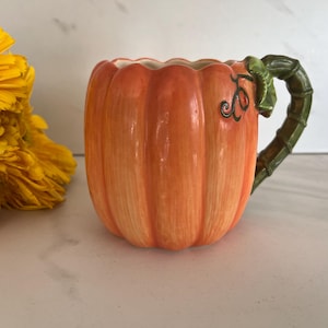 May include: A ceramic mug shaped like a pumpkin with a glossy orange finish and vertical ridges. The handle is designed to resemble a green, textured pumpkin vine with a small leaf detail attached to the side of the mug body.