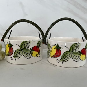 May include: Two white ceramic bowls with green handles and a strawberry design. The bowls have a white background with green leaves and red and yellow strawberries.