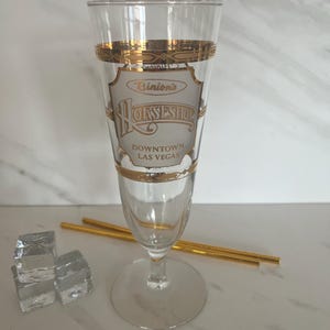 May include: A clear champagne flute with gold trim and the Binion's Horseshoe Downtown Las Vegas logo. The glass is next to three ice cubes and two gold-colored straws.