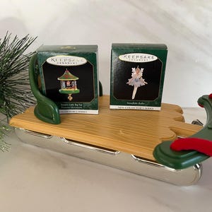 May include: Two small, green and white boxes with the words "Keepsake Ornament" on them. The boxes are sitting on a wooden sled with a red and green handle. The box on the left has a picture of a carousel with the words "Santa's Little Big Top" and "Victorian Movement" on it. The box on the right has a picture of a ballerina with the words "Snowflake Ballerina" on it. Both boxes have the words "New Collector's Series" on them.