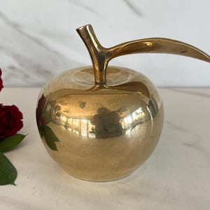 May include: A polished brass apple figurine with a long, curved stem. The surface of the metallic apple features a reflective gold finish and a small painted floral detail on the side, depicting red rose petals and green leaves.