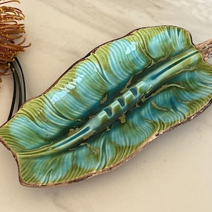 May include: A ceramic ashtray shaped like a large leaf with a glossy glaze in shades of turquoise and lime green. The piece features a central stem detail, textured veins, and a brown-toned edge, with integrated notches for resting items.