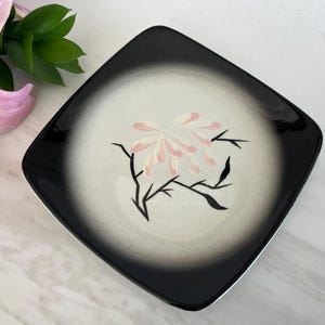 May include: Square black and white decorative plate with a floral design. The plate features a black border that fades into a white center. A pink and white flower design is in the center, with black stems and leaves.