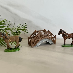 May include: Two brown horses with saddles and bridles stand on green bases. A wooden bridge with a gray stone archway is in the center.