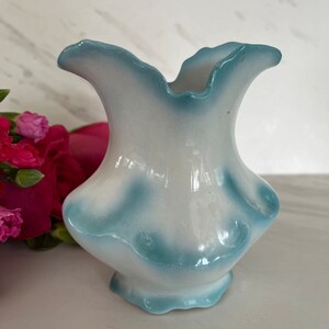 May include: A small, white ceramic vase with a blue glaze. The vase has a ruffled top and bottom.