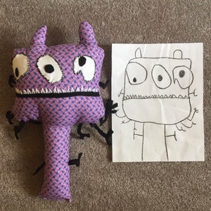 May include: A handmade purple monster plush toy with a patterned fabric body, large white eyes, and a black stitched mouth. Next to it is a child's drawing of a similar monster. The toy has black felt appendages.