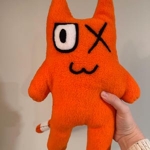 May include: Orange plush toy in the shape of a cartoon cat-like creature. The toy has a white and black eye, an X for an eye, and a stitched smile. It has small arms, legs, and a tail with white and black details.