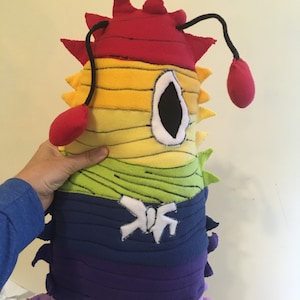 以下が含まれることがあります： 手作りの海の生き物型のぬいぐるみ。赤、黄、緑、青、紫など、虹色のグラデーションカラーが特徴です。大きな白い縁取りの目と、赤い球根状の端が付いた2本の黒い触角があります。