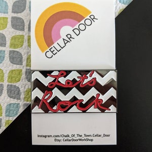 May include: Red enamel pin with the words "Let's Rock" in a white and black chevron pattern. The pin is on a white card with the text "Cellar Door" and social media information.