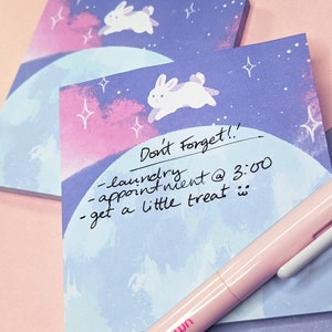 May include: A blue and pink notepad with a white bunny illustration and the text "Don't Forget!! - laundry - appointment @ 3:00 - get a little treat" written on it. A pink pen with a white bunny on the top is lying on the notepad.