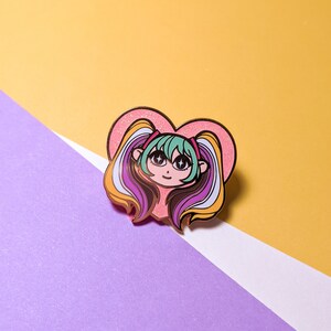 May include: A colorful enamel pin featuring a cartoon girl with large eyes and pigtails, set against a pink glitter heart. The girl has green hair and rainbow-colored pigtails. The pin is on a yellow and purple background.
