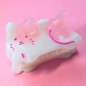 May include: A white and pink cat-shaped hair claw clip. The clip features a cat face design with pink accents for the ears, eyes, and mouth. The claw clip is made of a pearlescent material and is set against a pink background.