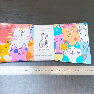 May include: A rectangular fabric item with a repeating pattern of cartoon cats in various colours. A metal ruler, marked in centimetres, is placed below the item. A tag with the text "Peridot Fox" is attached to the item.