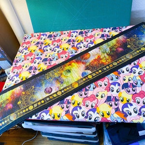 May include: A long, black fabric table runner with a colorful galaxy print and gold accents. The runner is laid on a table covered in a white fabric with a repeating pattern of colorful cartoon pony faces.
