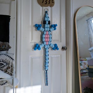 May include: A beaded lizard decoration in shades of blue, pink, and black hangs on a white door. The lizard has a long tail and is made of round beads. A small, brown, cloud-shaped item with the number "133=" is above the lizard.