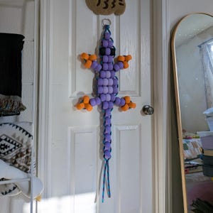 May include: A beaded lizard decoration in shades of purple, orange, and black hangs on a white door. The lizard has a long tail and four limbs. The door has a doorknob and a small, woven sign above it.