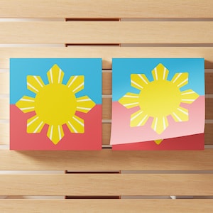 May include: Two square prints featuring a yellow sun with eight rays against a split background of light blue on top and red on the bottom. The right print shows a corner folded over, revealing a light pink layer underneath the top surface.