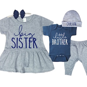 May include: A gray and blue sibling outfit set for a baby boy and girl. The girl's outfit includes a gray dress with a blue "big sister" graphic and a blue bow headband. The boy's outfit includes a blue bodysuit with a white "little brother" graphic, gray pants, and a gray hat with the name "CARSON" embroidered in blue.