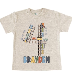 May include: A light gray t-shirt with a colorful train design forming the number four. The shirt has the name "BRAYDEN" printed in colorful letters below the train design.