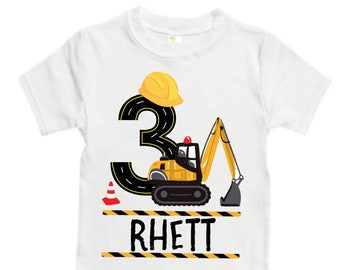 May include: White t-shirt with a yellow construction vehicle and the number 3. The vehicle has a yellow hard hat on top of the number. The text "RHETT" is printed below the vehicle.