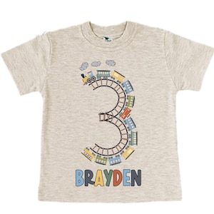 May include: A light grey t-shirt with a graphic of a train going around the number 3. The name "BRAYDEN" is printed below the number.