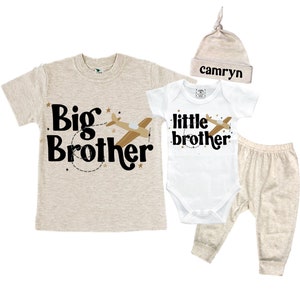 May include: A beige and white baby outfit set with a beige hat. The shirt says "Big Brother" with a plane and stars. The bodysuit says "little brother" with a plane and stars. The hat says "camryn".