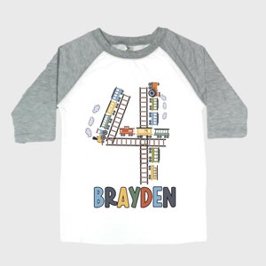 May include: White and gray raglan-style shirt with a colorful train-themed design. The number "4" is formed by train cars and tracks. The name "BRAYDEN" is printed below the number in blue, orange, and green.