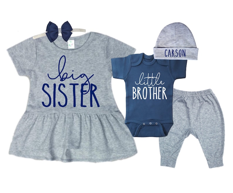 May include: A gray and blue sibling outfit set for a baby boy and girl. The girl's outfit includes a gray dress with a blue "big sister" graphic and a blue bow headband. The boy's outfit includes a blue bodysuit with a white "little brother" graphic, gray pants, and a gray hat with the name "CARSON" embroidered in blue.