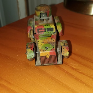 May include: A vintage toy tank with a camouflage pattern in shades of green, brown, and yellow. The toy has a detailed design with visible wheels, a turret, and a front grill. The toy is made of metal and has a weathered appearance.