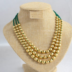 Puede incluir: Un collar de cuentas doradas y verdes con tres hileras de cuentas doradas grandes y redondas y dos hileras de cuentas verdes más pequeñas.