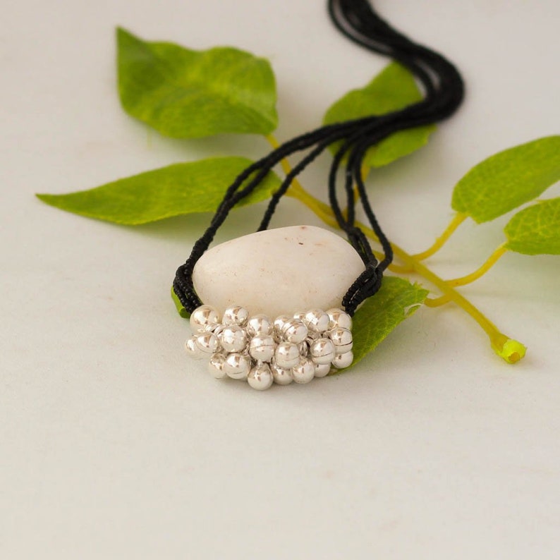 May include: A silver charm necklace with a cluster of small round beads. The necklace is on a black cord and is resting on a white stone.