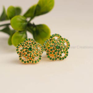 May include: A pair of gold-colored flower-shaped stud earrings. Each earring is encrusted with small, round, emerald-green gemstones. The earrings are set against a soft, neutral background with a blurred green plant in the background.