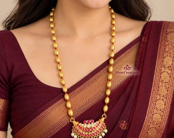 May include: A long gold-toned necklace featuring oval-shaped beads separated by small red accents. The pendant is a crescent-shaped piece adorned with multiple rows of small pink stones and a bottom fringe of tiny white pearls. The text ZivarCreations is visible.