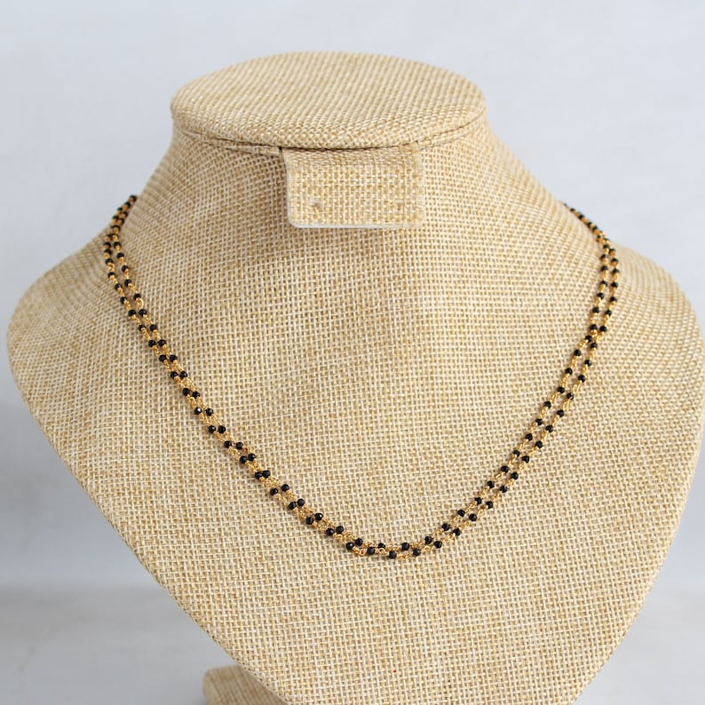 May include: A gold chain necklace with black beads. The necklace is displayed on a tan fabric mannequin.