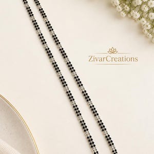 Puede incluir: Un collar de doble cadena con pequeñas cuentas negras alternadas con cuentas metálicas de color plateado. El collar incluye un cierre de mosquetón en cada extremo y muestra el texto ZivarCreations con un icono de loto impreso.