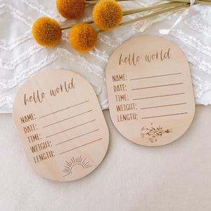 May include: Two wooden baby announcement plaques with the words "hello world" in script. Each plaque has lines for name, date, time, weight, and length. One plaque has a sun design, the other has a floral design.