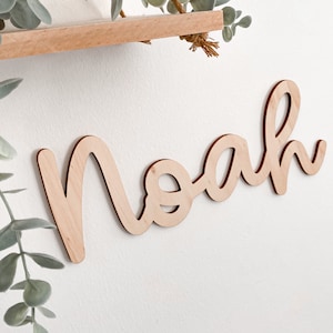 May include: Wooden name sign with the name "Noah" written in a cursive font. The sign is made of light brown wood and is hanging on a white wall.