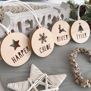 May include: Four wooden Christmas baubles with different designs and names. The baubles are round and have a white string attached to them. The names on the baubles are Harper, Chloe, River, and Tyler. The designs on the baubles are a star, a snowflake, a reindeer, and a Christmas tree.