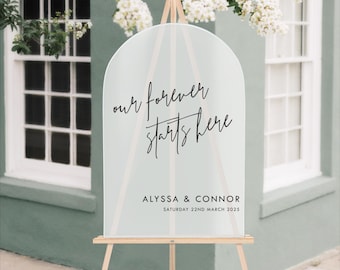 May include: A white acrylic wedding sign with a black arch shape and the text "our forever starts here" and "Alyssa & Connor Saturday 22nd March 2025".