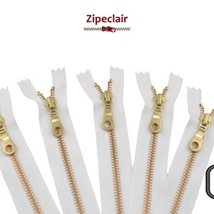 Peut inclure: Cinq fermetures éclair en métal doré avec un ruban blanc. Les fermetures éclair sont ouvertes et le curseur est au milieu de la fermeture éclair. Le texte "METAL DORE" est dans le coin inférieur gauche de l'image. Le texte "501 BLANC" est dans le coin inférieur droit de l'image.