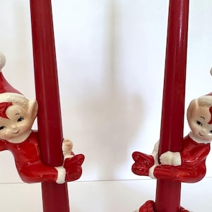 May include: A pair of ceramic candle holders featuring two identical elf figures dressed in red suits with white trim and matching red hats with white pom-poms. Each elf is depicted climbing a tall, smooth, solid red taper candle.