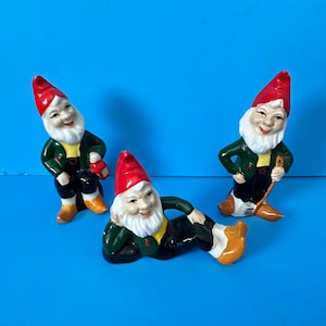 May include: Three ceramic gnomes with red hats and white beards. One gnome is holding a lantern, another is holding a broom, and the third is lying down.