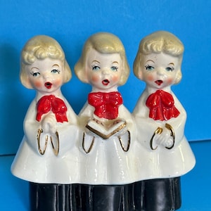 May include: Three porcelain figurines of children singing in white robes with red bows and black pants. The middle child is holding a book.