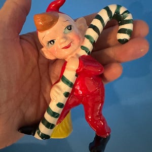 May include: A vintage ceramic Christmas elf figurine. The elf is wearing a red outfit, black shoes, and a red hat. It is holding a green and white striped candy cane. The elf has a smiling face with rosy cheeks and a yellow bag.
