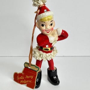 May include: A vintage Christmas ornament depicting a smiling elf in a red Santa suit with a white trim. The elf has blonde hair and black boots. A tag that says "Santa's Merry Helpers" hangs from the ornament.