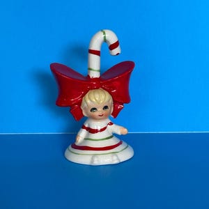 May include: A ceramic Christmas figurine featuring a girl with blonde hair, wearing a white dress with red and green stripes. A red bow is behind her head, and a candy cane arches above. The figurine is on a white base.