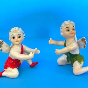 May include: Two ceramic cherub figurines with white skin, gold-tipped wings, and detailed facial features. One wears a red outfit, the other a green outfit. Both are kneeling on a blue surface.