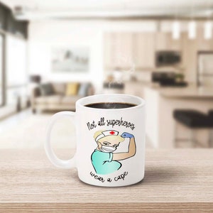 Peut inclure: Mug en c&eacute;ramique blanche remplie de caf&eacute; noir, avec une infirmi&egrave;re de dessin anim&eacute; montrant ses muscles. Le mug porte l'inscription "Not all superheroes wear a cape". L'infirmi&egrave;re porte un masque et une coiffe d'infirmi&egrave;re.